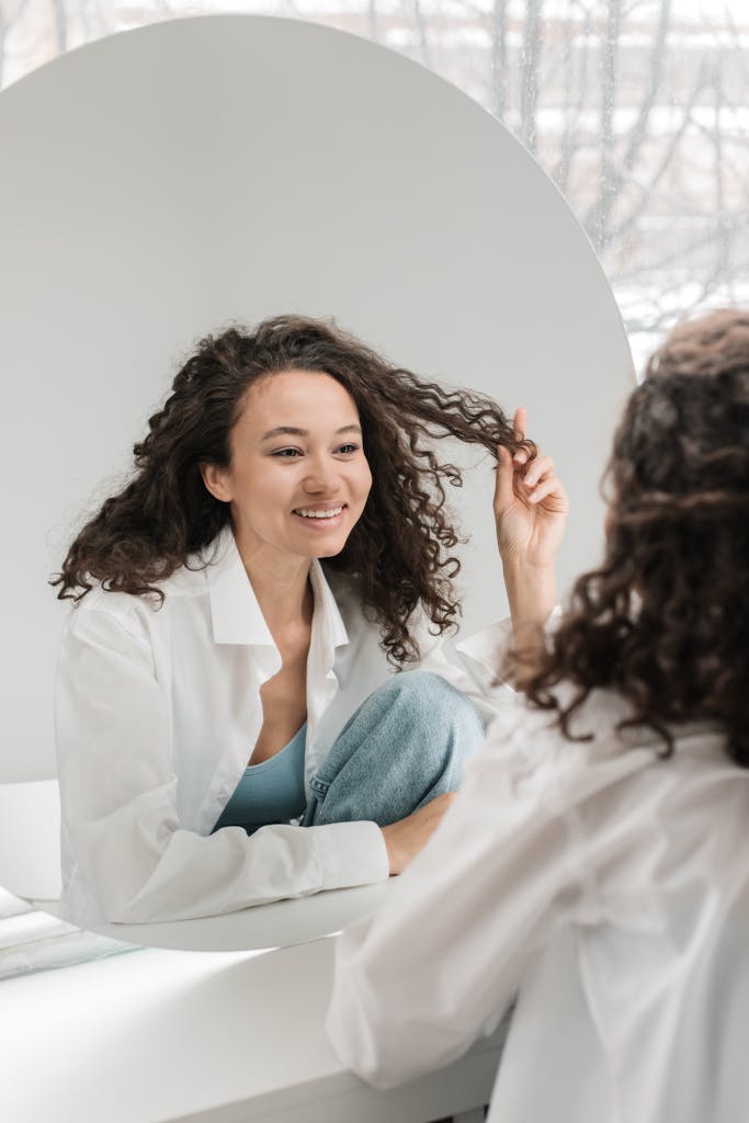 Happy woman with curly hair smiling at her reflection in a mirror, embracing natural beauty.