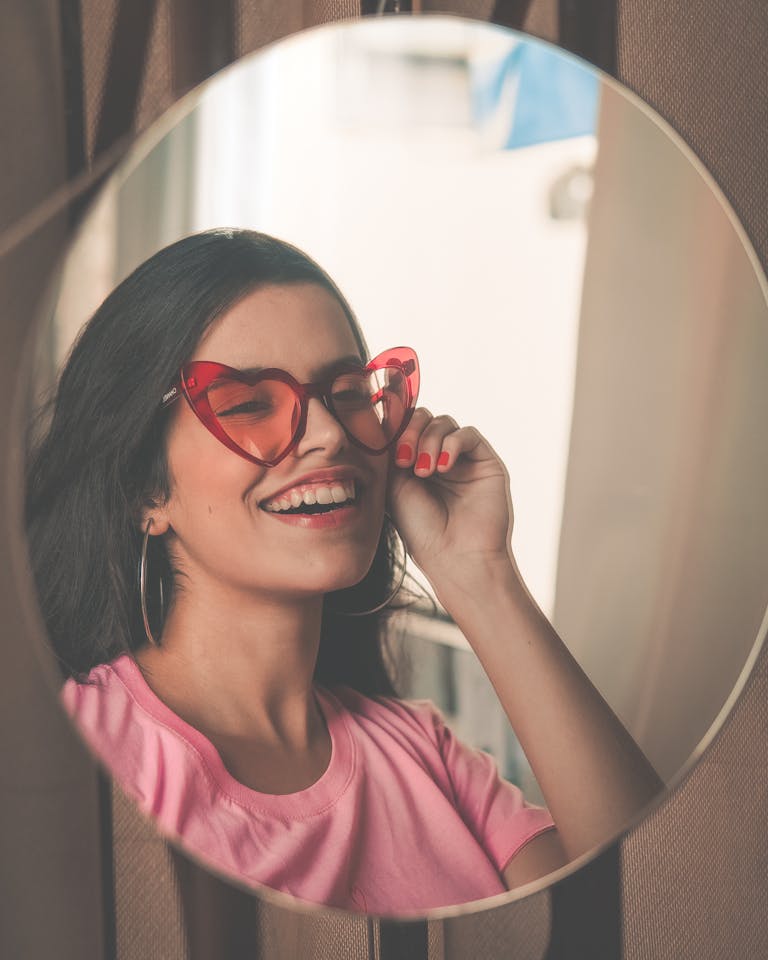 Stylish woman wearing heart-shaped sunglasses smiles joyfully at her reflection in a mirror with the goal on how to validate yourself.