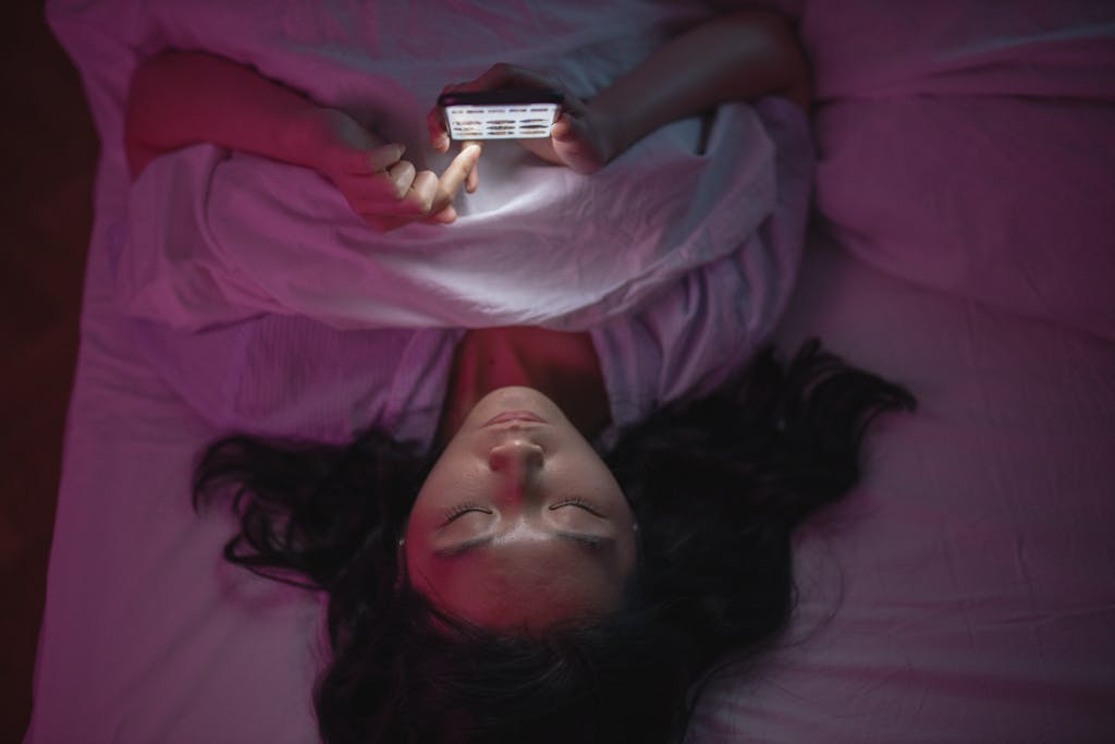 A woman lying in bed at night doomscrolling her smartphone under soft lighting.